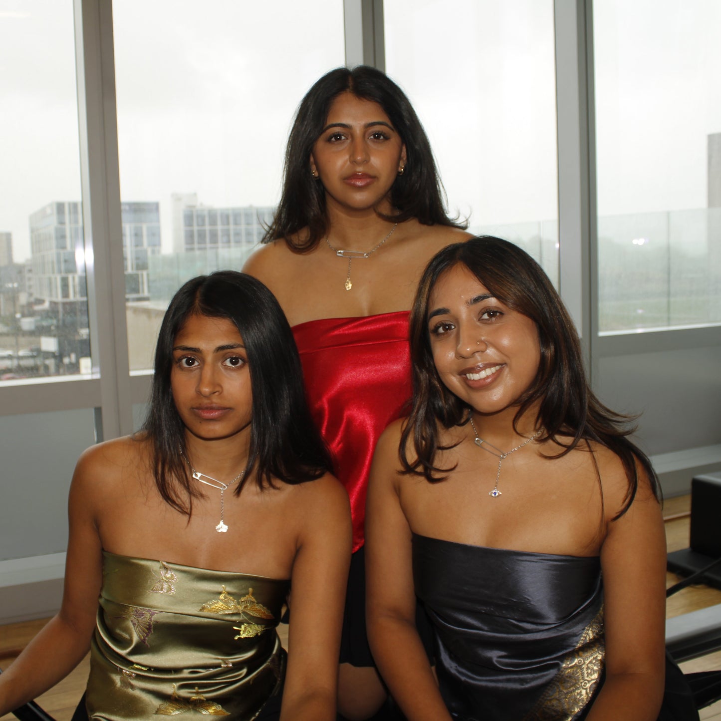 Three women in strapless dresses posing indoors with a cityscape view through large windows.