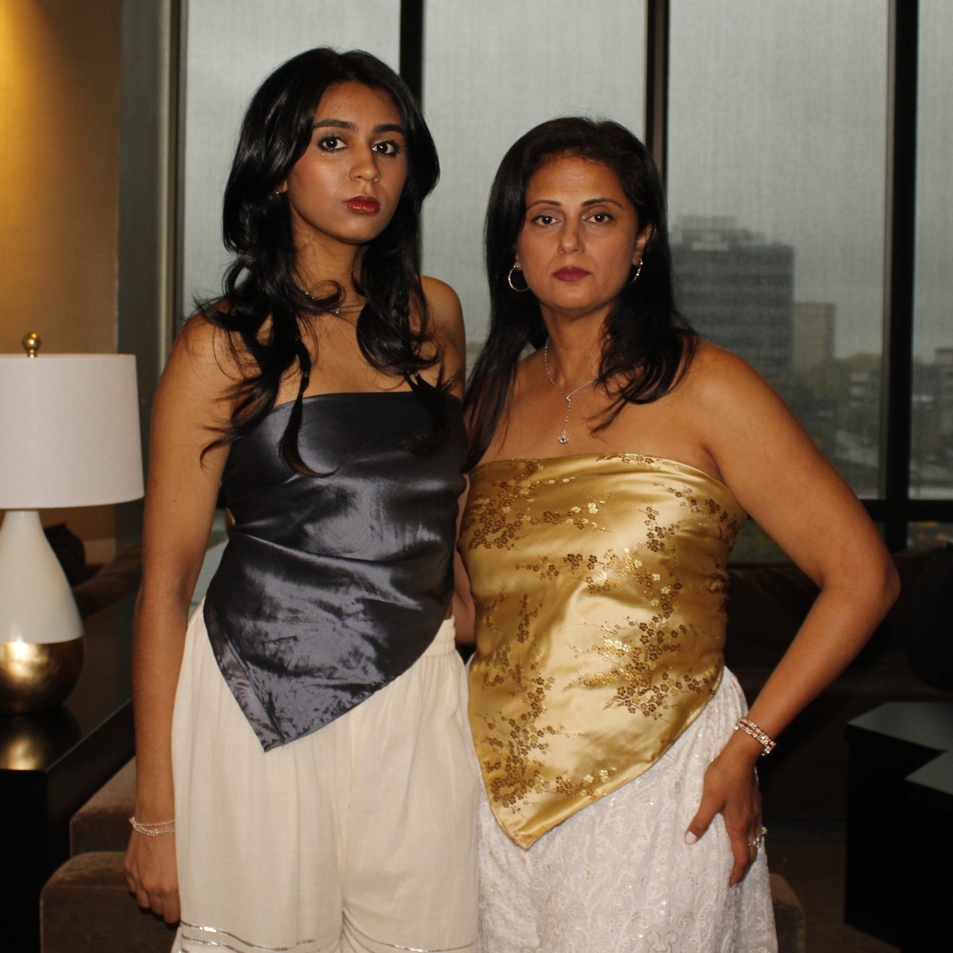 Two women in formal attire standing in a room with large windows showing an urban landscape.